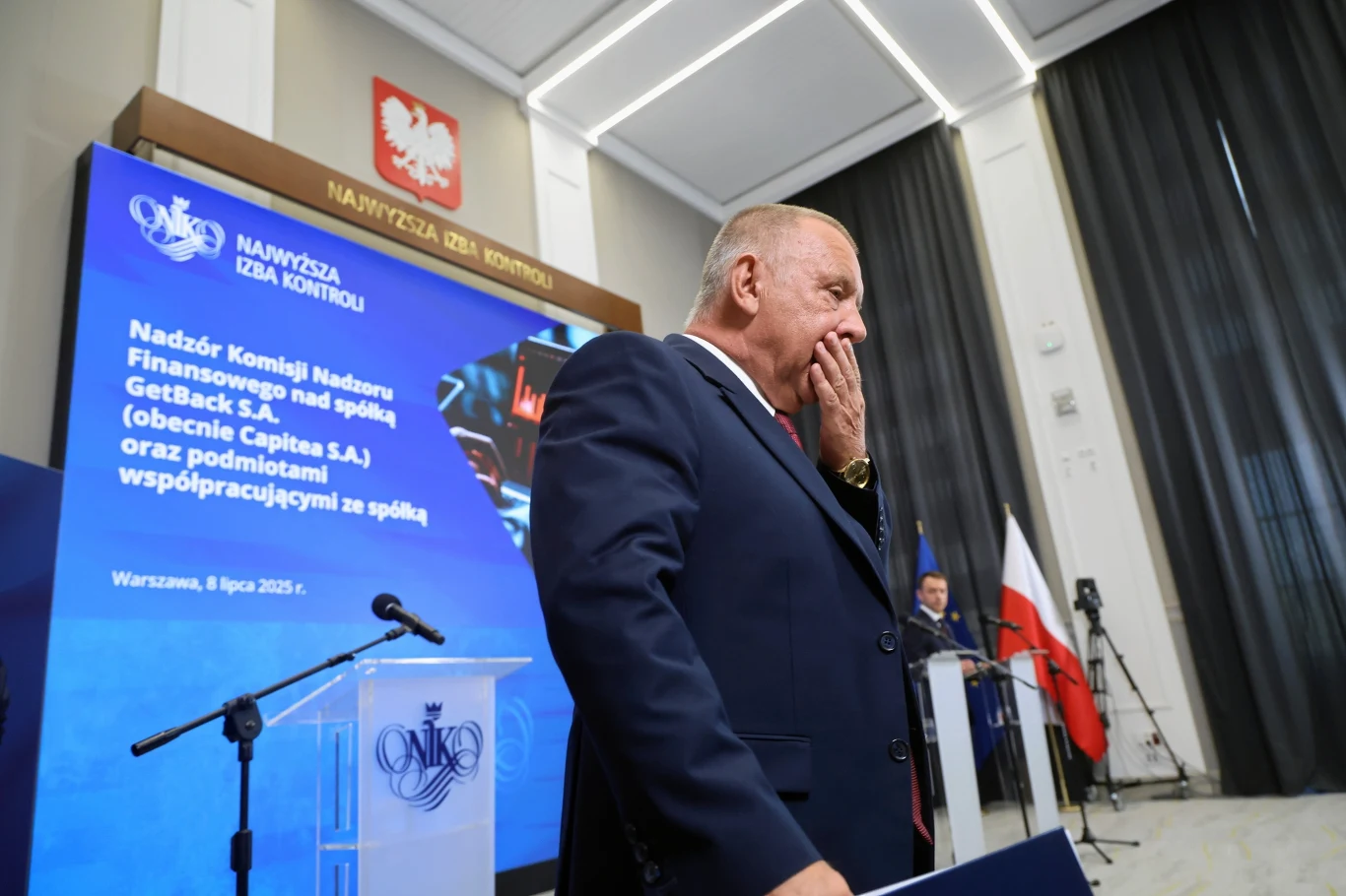 Starszy mężczyzna w garniturze stoi na tle podestu z mikrofonami i niebieskim ekranem informacyjnym Najwyższej Izby Kontroli. Za nim znajduje się godło Polski oraz polska flaga. W tle kolejny mężczyzna na scenie.