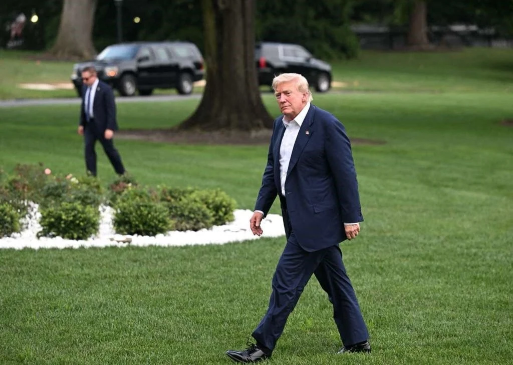 Prezydent USA Donald Trump Mężczyzna w granatowym garniturze idzie po trawniku, w tle widoczny jest drugi mężczyzna oraz czarne samochody zaparkowane przy drzewach.
