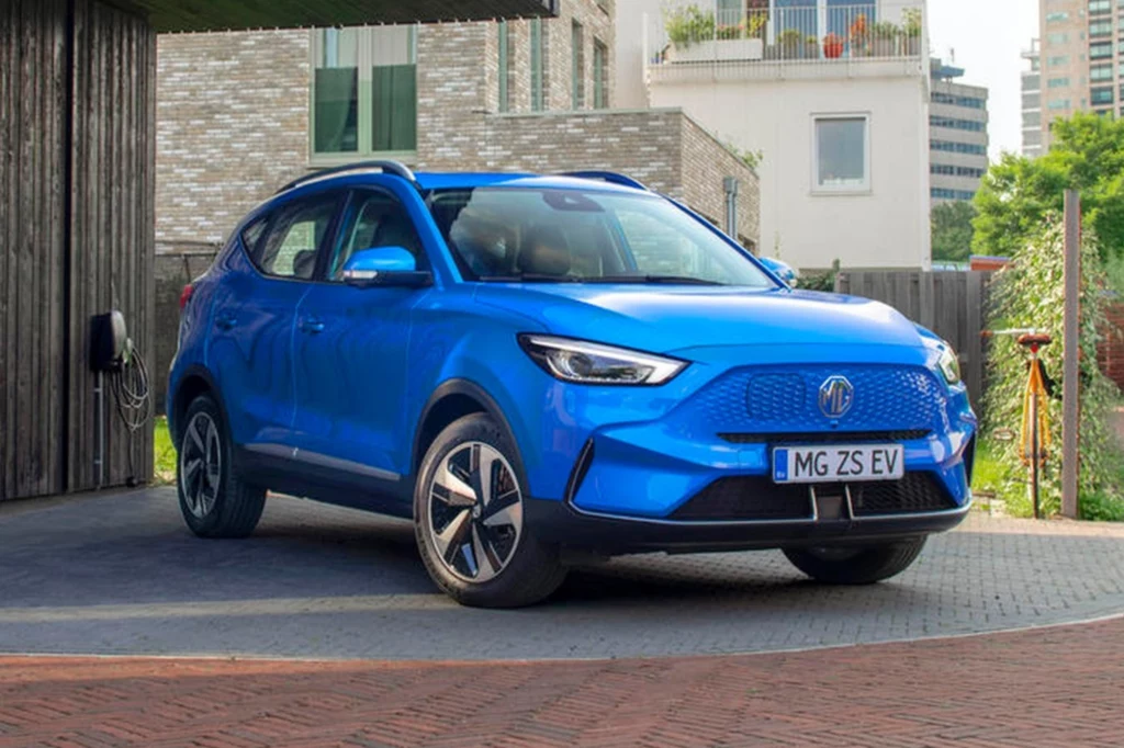 Cena tego modelu na rynku brytyjskim rozpoczyna się od 29 770 funtów. Niebieski elektryczny samochód MG ZS EV stoi zaparkowany przy stacji ładowania na tle nowoczesnej zabudowy miejskiej, pod lekkim cieniem budynku.