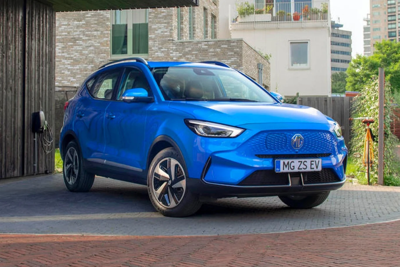 Niebieski elektryczny samochód MG ZS EV stoi zaparkowany przy stacji ładowania na tle nowoczesnej zabudowy miejskiej, pod lekkim cieniem budynku.