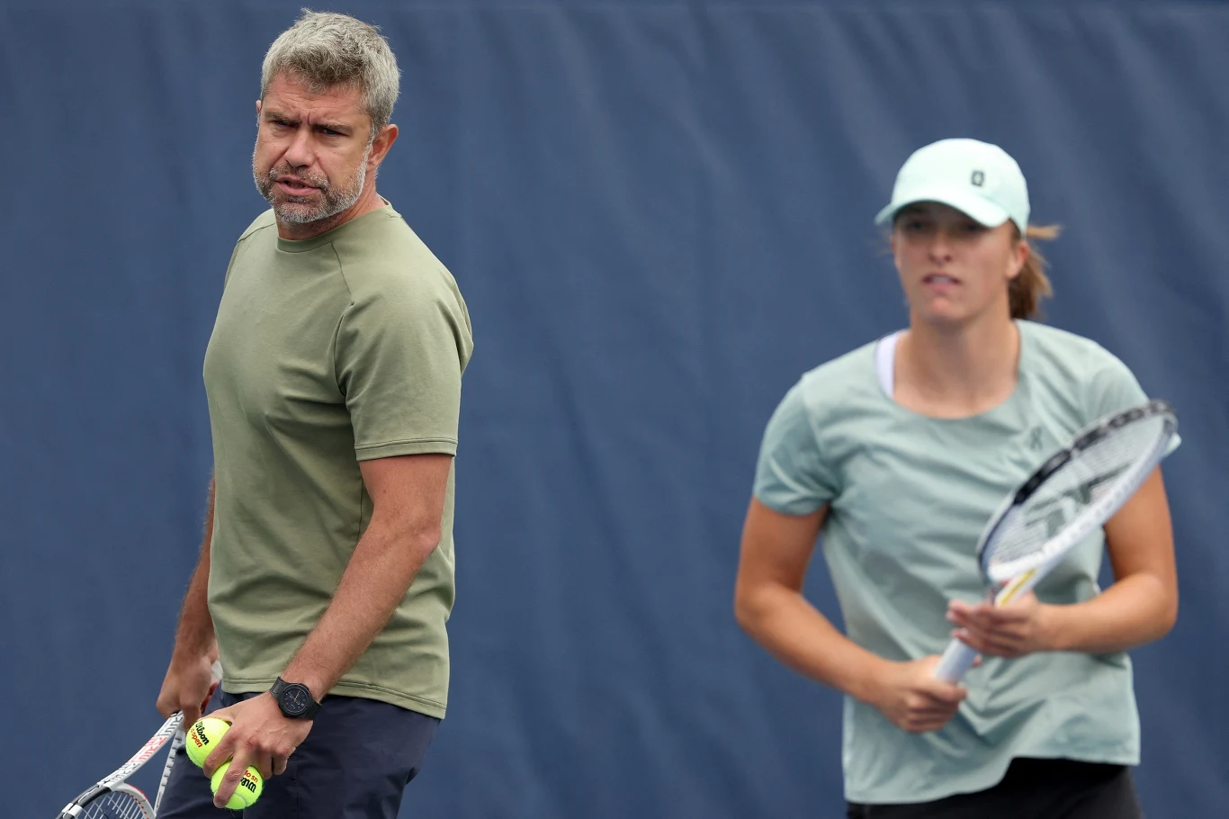 Dwoje ludzi na tle niebieskiego kortu tenisowego, mężczyzna w zielonej koszulce trzyma dwie piłki tenisowe oraz rakietę, kobieta w niebieskiej koszulce i czapce trzyma rakietę tenisową