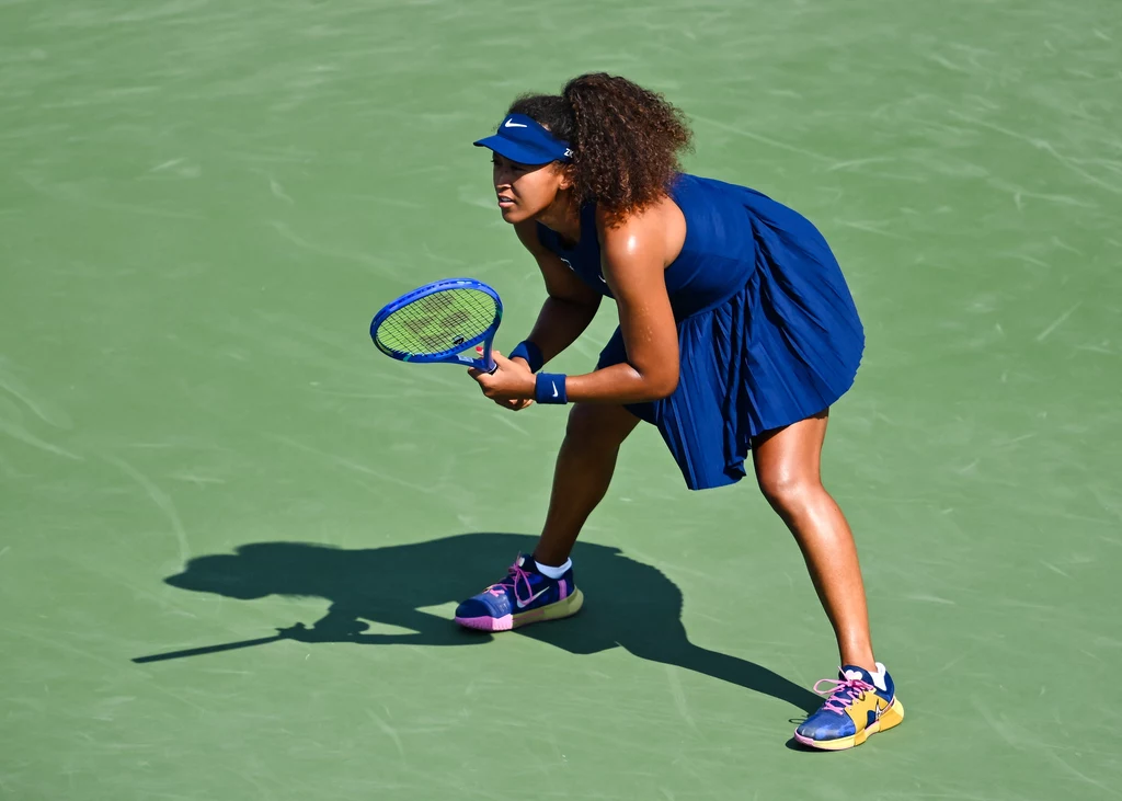 Naomi Osaka Tenisistka w ciemnoniebieskiej sukience i sportowym obuwiu przygotowuje się do odbioru serwisu na zielonym korcie, skupiona na grze, z rakietą w ręce i lekko ugiętymi nogami.
