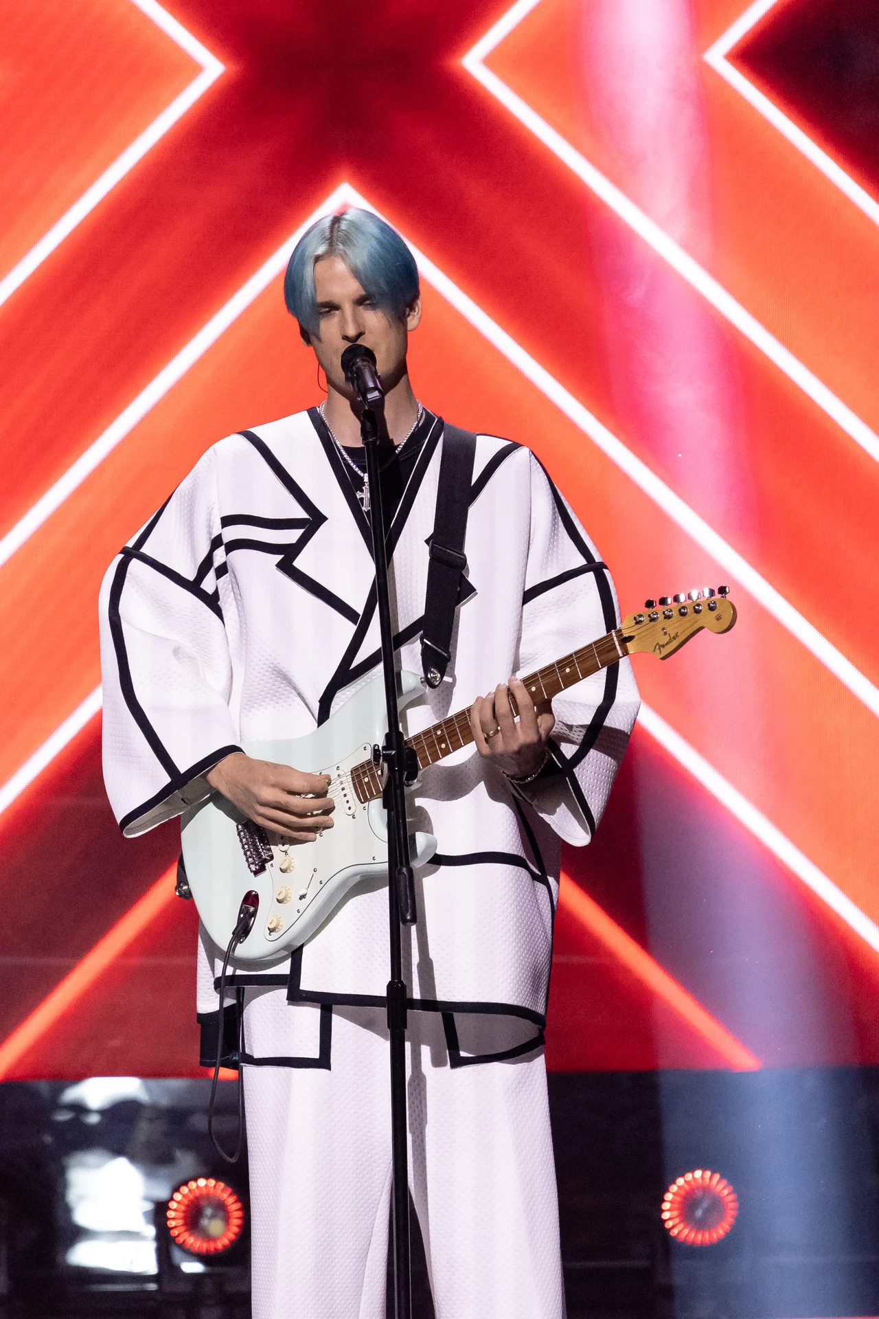Muzyk z niebieskimi włosami ubrany w biały strój z czarnym wzorem gra na elektrycznej gitarze na tle czerwonej scenografii z wyrazistymi, świetlnymi kształtami w formie litery X.