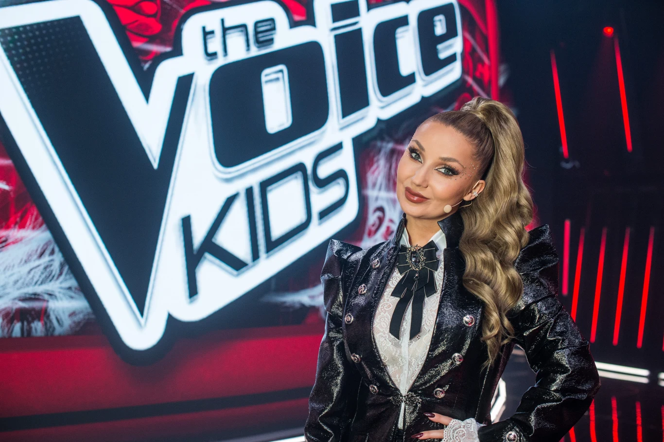 Cleo została trenerką dziewiątej edycji "The Voice Kids". Kto zasiądzie u jej boku? Kobieta w eleganckim, czarnym stroju z kokardą, stojąca na tle dużego, podświetlonego logotypu programu The Voice Kids, scena utrzymana w czerwono-czarnej kolorystyce.