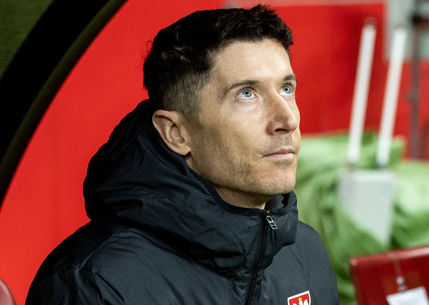 Mężczyzna w czarnej kurtce sportowej siedzi na ławce przy boisku, patrzy w górę w zamyśleniu, w tle czerwone elementy stadionowe.