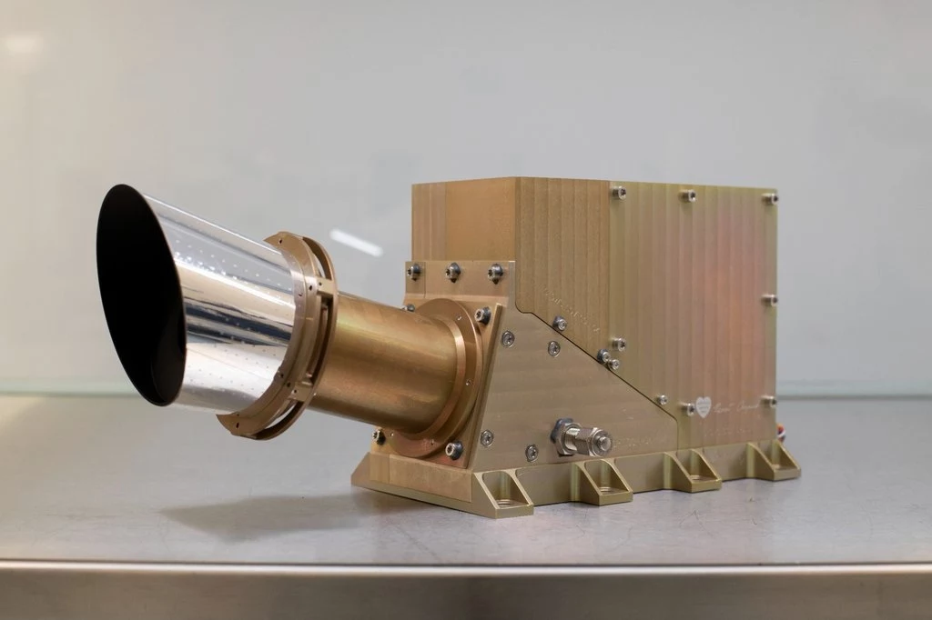 Polski instrument GLOWS dla sondy NASA IMAP, który stworzono w CBK PAN. Metaliczne urządzenie o prostokątnym korpusie z wystającą stożkową tubą, stojące na gładkiej powierzchni stołu, w tle jednolita jasna ściana.