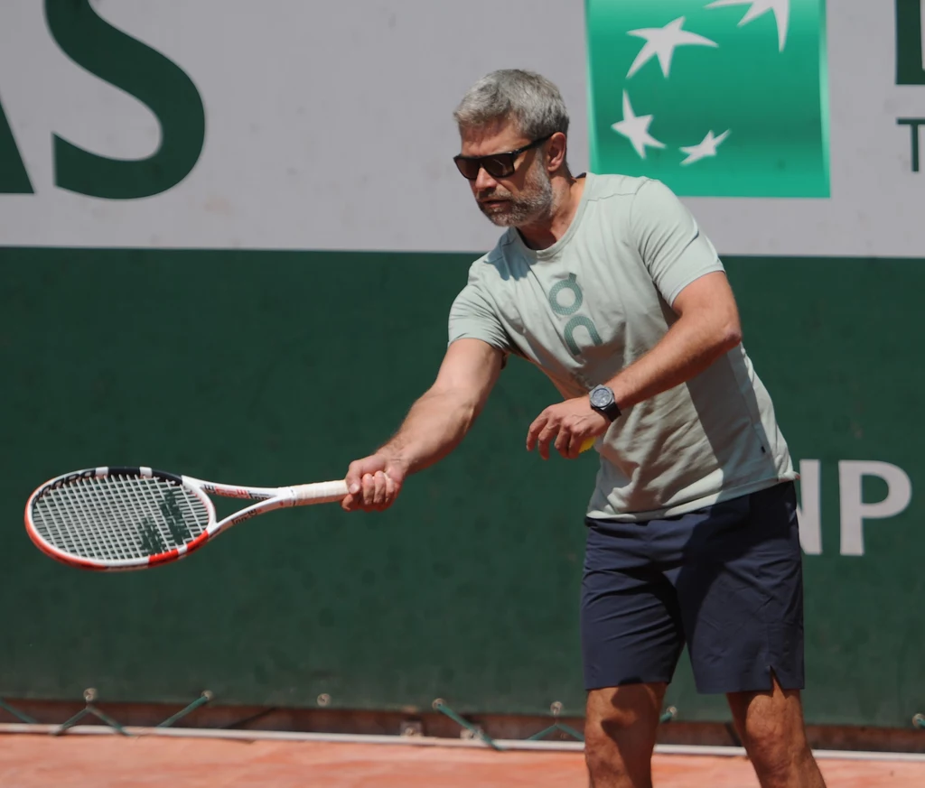 Tomasz Wiktorowski Mężczyzna w okularach przeciwsłonecznych i sportowym stroju gra w tenisa na korcie ziemnym, trzyma rakietę w pozycji gotowości do odbicia piłki.