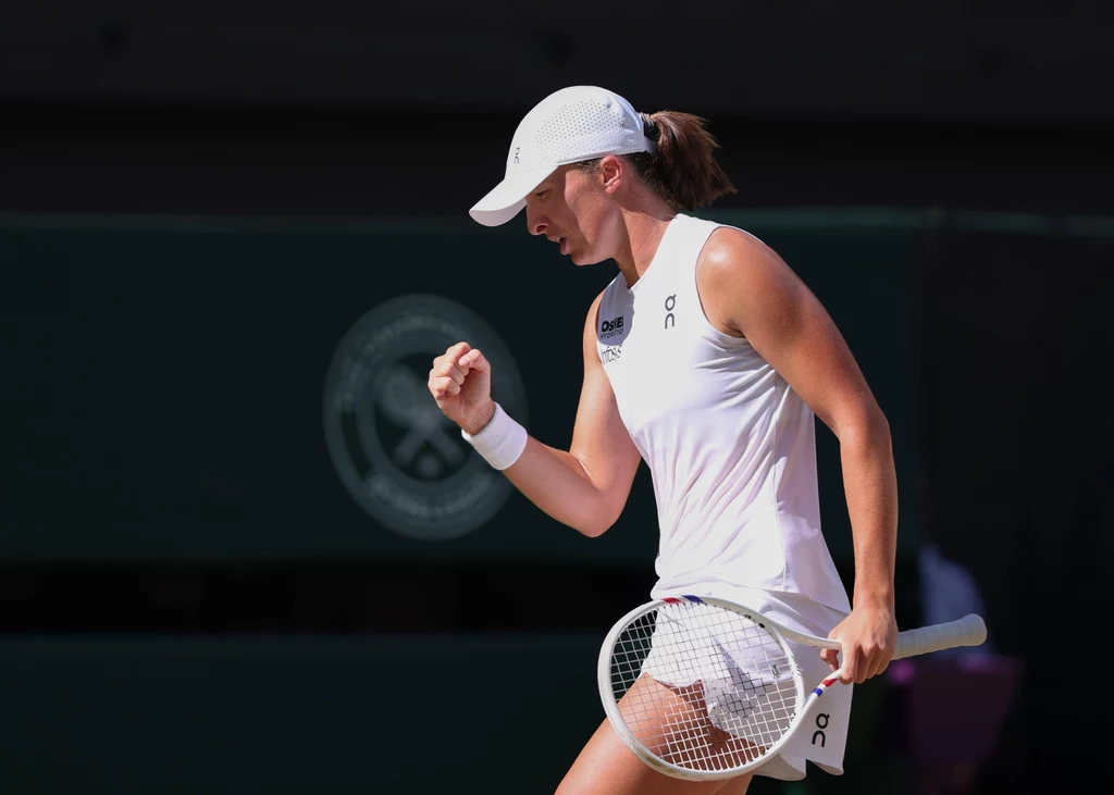 Iga Świątek Tenisistka ubrana na biało wykonuje gest zwycięstwa, trzymając rakietę tenisową na tle kortu z widocznym symbolem turnieju Wimbledon.