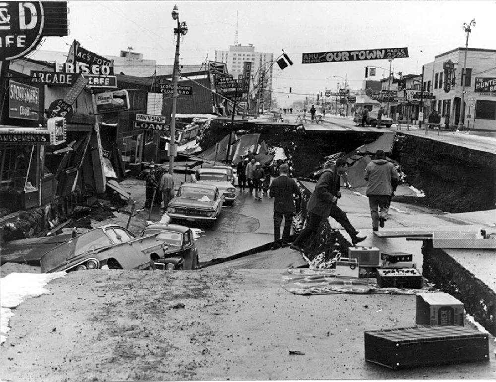 Trzęsienie ziemi na Alasce w 1964 roku Zniszczona ulica miejska z zapadniętym asfaltem i uszkodzonymi budynkami, grupa ludzi obserwuje skutki katastrofy, na drodze stoją samochody, część z nich została uszkodzona przez zapadlisko.