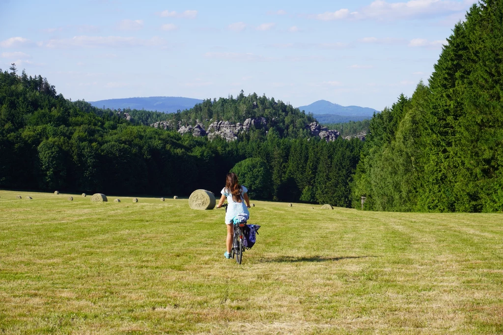 Szlak wokół Skalnych Miast zachwyca przestrzeniami i panoramicznymi widokami Dziewczynka jadąca rowerem przez szeroką, otwartą łąkę, wokół baloty siana, w tle rozciągający się krajobraz lasu, wzgórz oraz odległych gór pod błękitnym niebem.