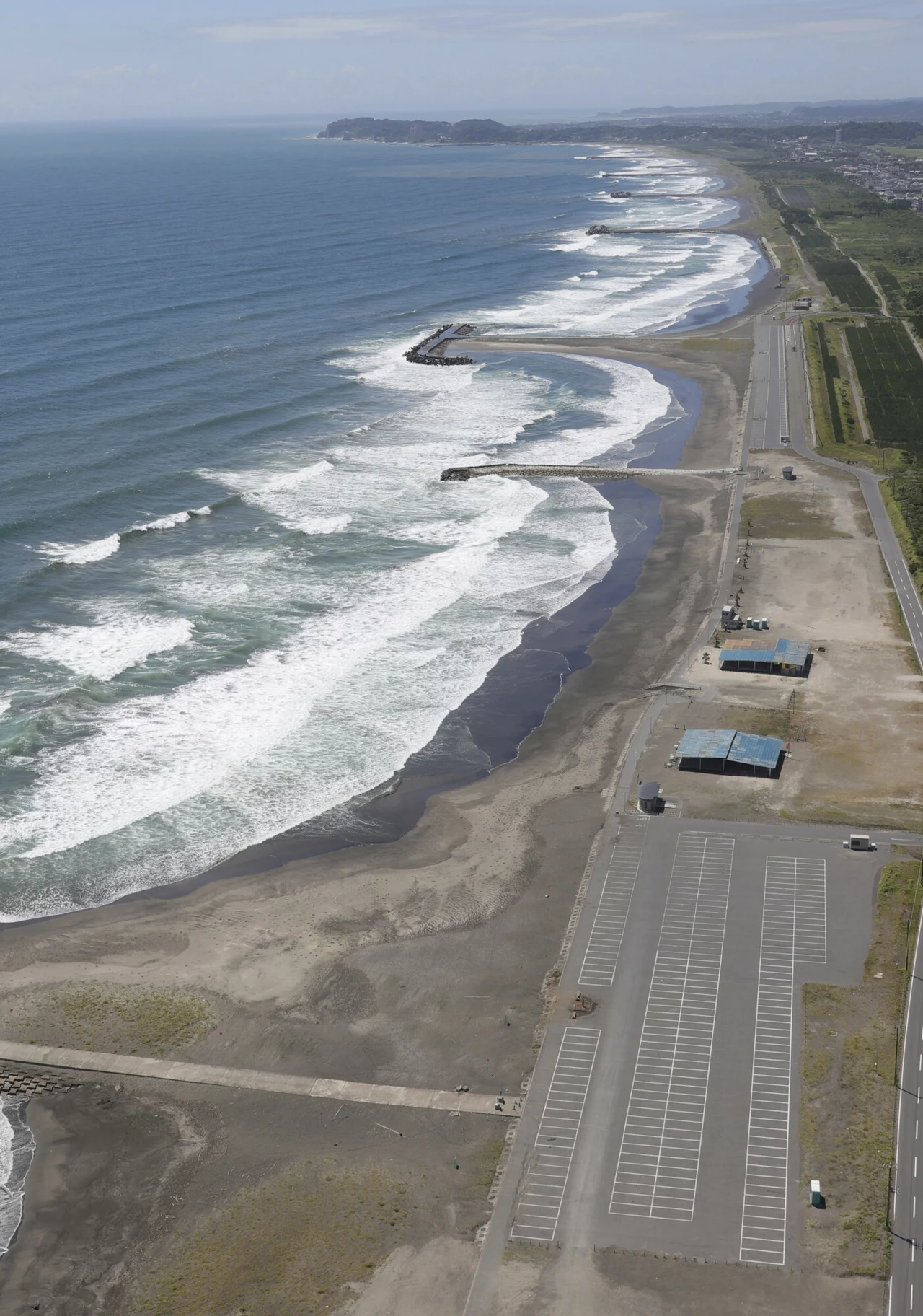 Fala tsunami powstaje po silnych trzęsieniach ziemi Linia wybrzeża z rozległą plażą, falami rozbijającymi się o brzeg, szerokim pasem asfaltowej drogi oraz dużym pustym parkingiem równolegle biegnącym wzdłuż morza, na horyzoncie pagórkowata linia lądu.