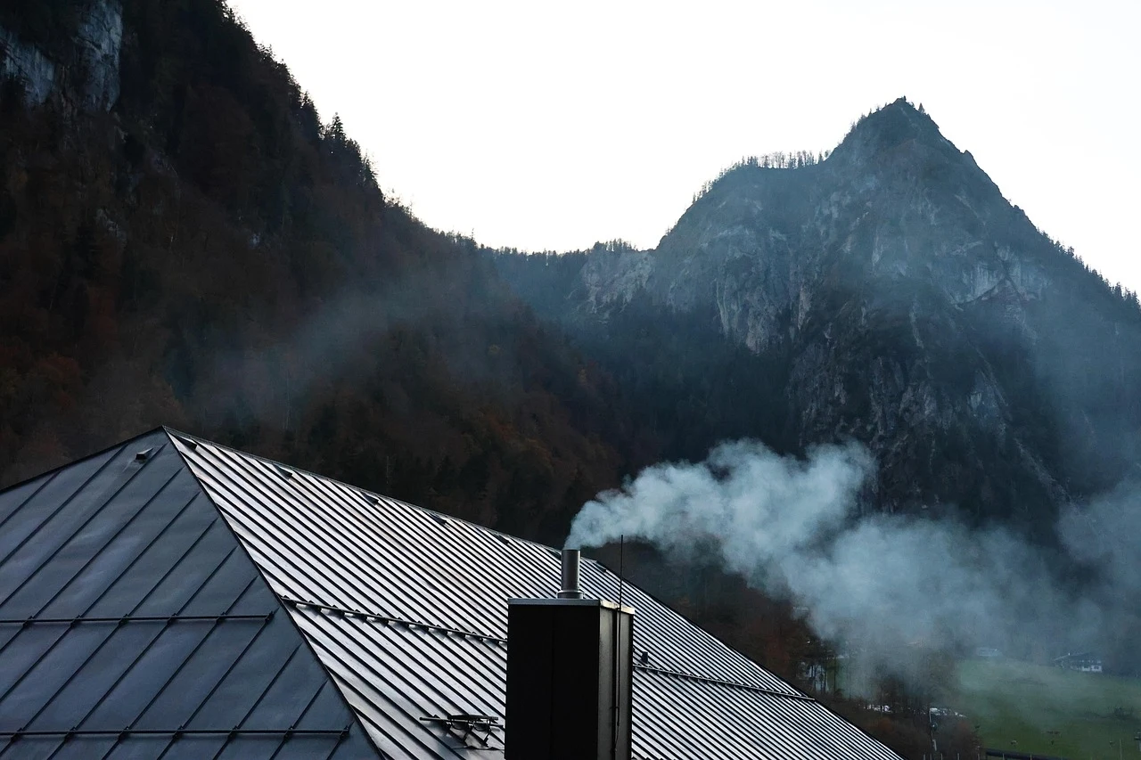 Dach budynku z metalowymi panelami oraz kominem, z którego unosi się biały dym na tle gór porośniętych lasem i szczytów w oddali.