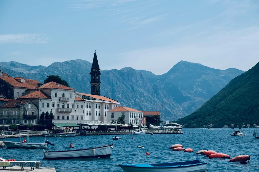 Malownicze miasteczko nad brzegiem zatoki, otoczone wysokimi górami, z charakterystyczną wieżą kościoła w tle i licznymi łodziami zacumowanymi przy brzegu.