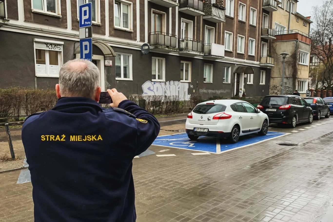 Jaki mandat grozi za nieuprawnione parkowanie na kopercie? Funkcjonariusz Straży Miejskiej wykonuje zdjęcie samochodu zaparkowanego na miejscu dla osób niepełnosprawnych obok innych pojazdów, w tle widoczne są bloki mieszkalne i znak parkingowy.