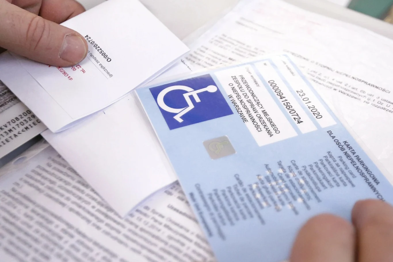 Aby uzyskać kartę inwalidy, należy złożyć odpowiedni wniosek. Ręce osoby trzymającej niebieską kartę parkingową osoby niepełnosprawnej oraz kilka dokumentów urzędowych.