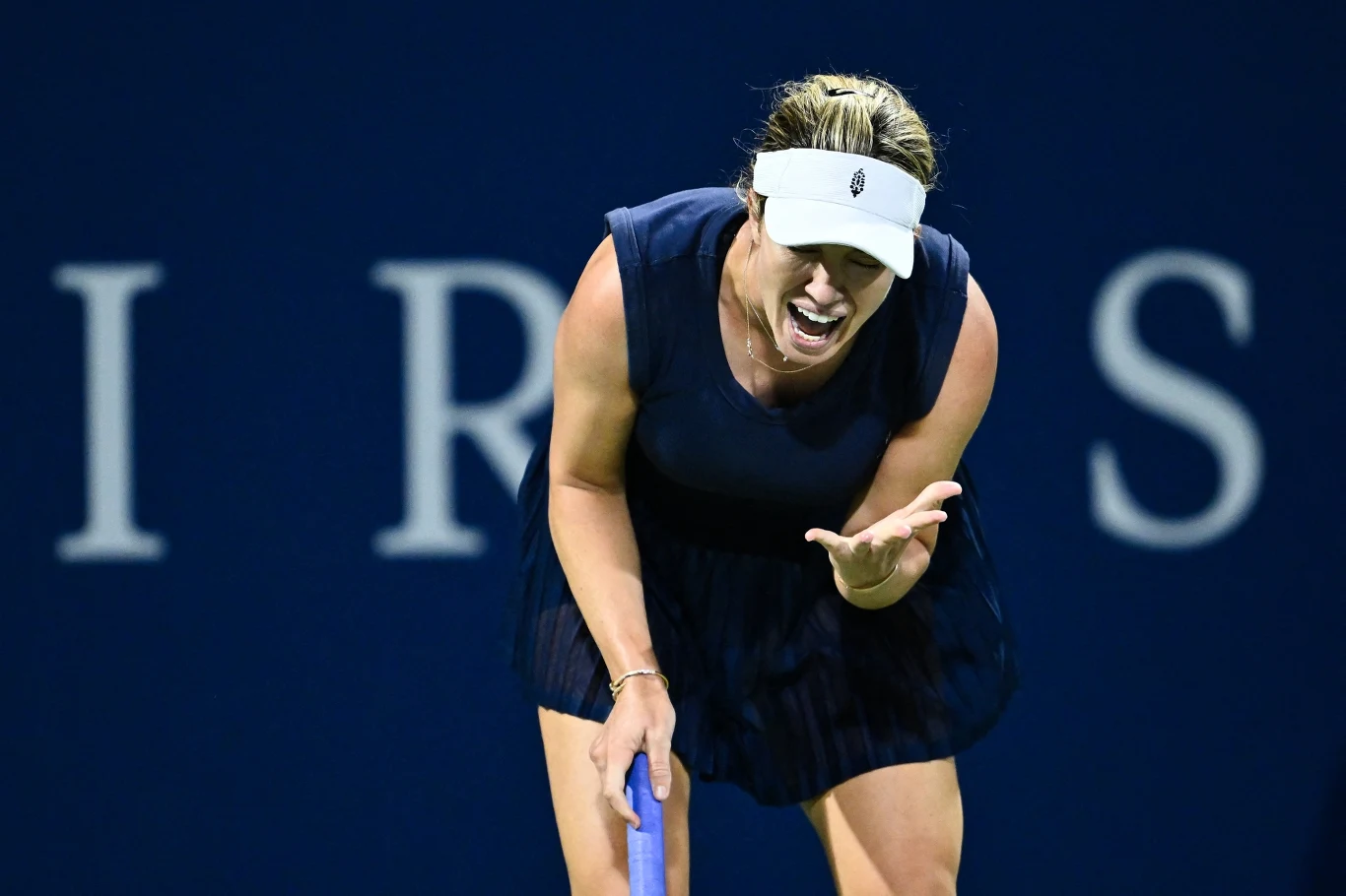 Danielle Collins Tenisistka w ciemnej sukience sportowej oraz białym daszku wyraża silne emocje podczas meczu tenisowego, pochylona do przodu, z widoczną frustracją na twarzy, na tle niebieskiego kortu.
