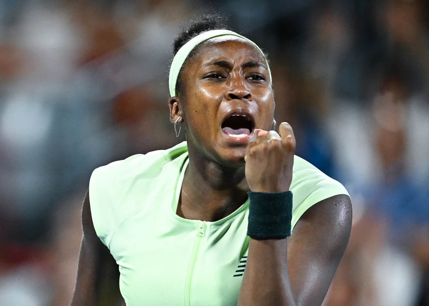 Kobieta w sportowym stroju, z opaską na głowie, zaciska pięść z wyrazem silnych emocji na twarzy, wyrażając triumf lub determinację podczas meczu tenisowego.