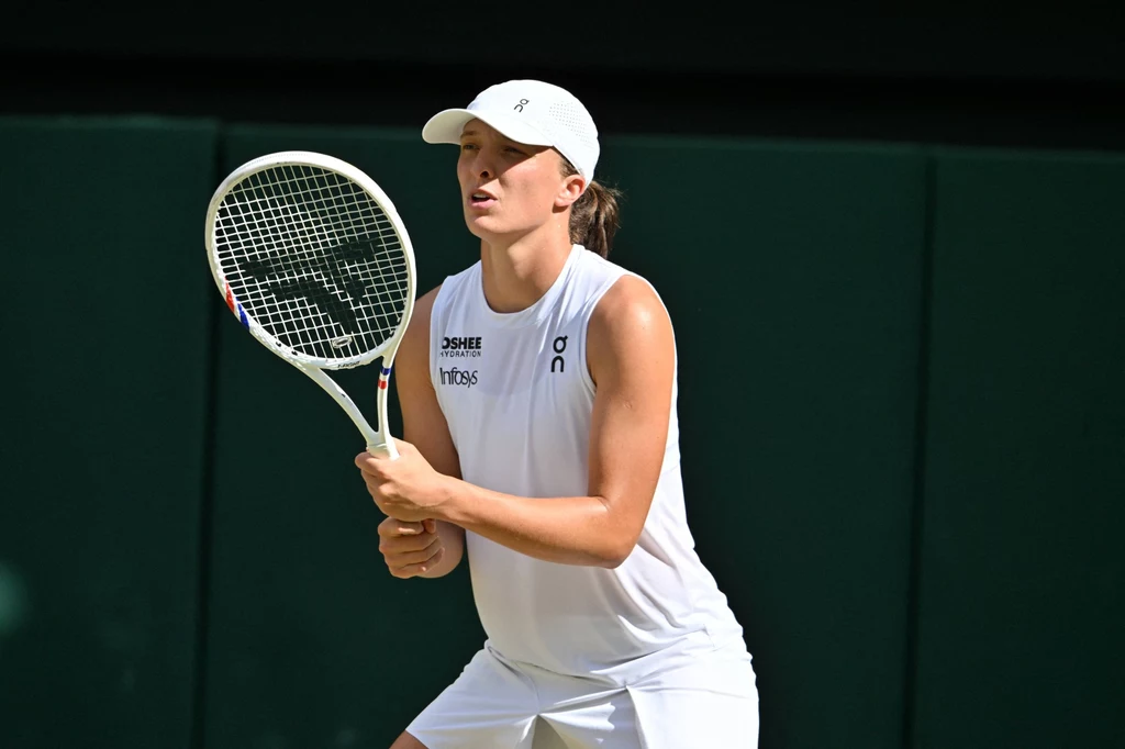 Iga Świątek Sportowiec ubrany na biało skupia się na akcji podczas gry w tenisa, trzyma rakietę w obu rękach i przygotowuje się do zagrania piłki; tło stanowi zielony kort.