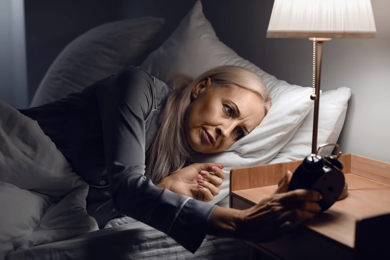 Pełnia Księżyca uderza w sen. Co drugi senior może to odczuć Starsza kobieta sięga po budzik na szafce nocnej, leżąc w łóżku oświetlonym lampką przy pełni Księżyca.