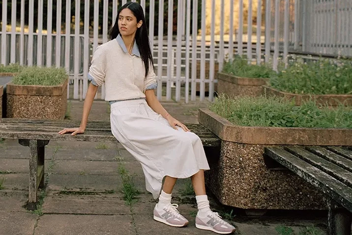 Sneakersy New Balance Młoda kobieta siedzi na drewnianej ławce w miejskim parku, ubrana w białą spódnicę, jasny sweter i sportowe buty, w tle betonowe donice z zielenią oraz metalowe ogrodzenie.
