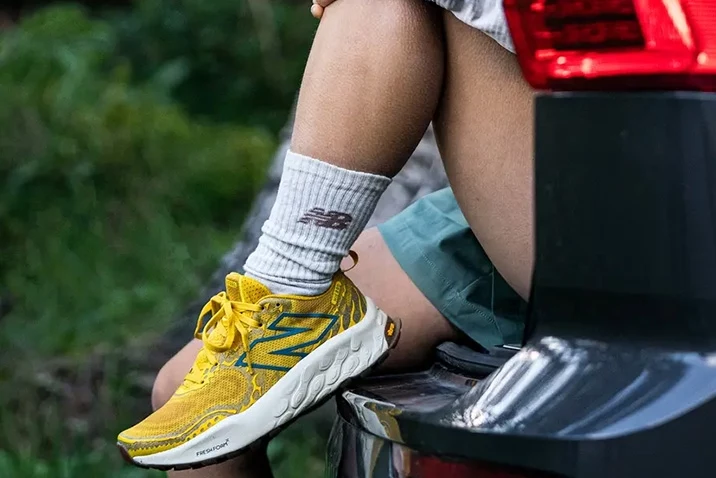 NB Fresh Foam X Hierro Żółte buty sportowe marki New Balance na nogach osoby siedzącej na tylnej klapie auta, jasne skarpetki z logotypem marki oraz fragmenty samochodu i zielonego otoczenia w tle.