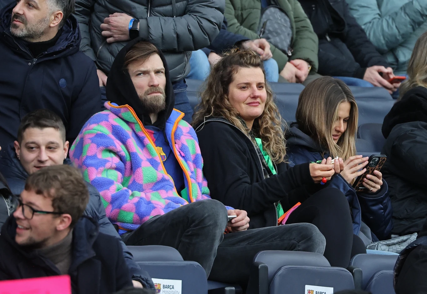 Grupa ludzi siedzących na widowni stadionu, skoncentrowana na wydarzeniu, kilka osób patrzy przed siebie, a jedna kobieta korzysta z telefonu, wśród widzów mężczyzna w kolorowej kurtce przyciąga uwagę.