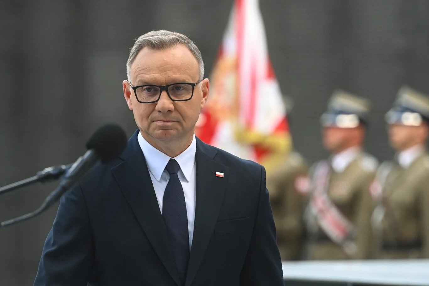 Mężczyzna w garniturze i okularach stoi przed mikrofonem, w tle rozmazani żołnierze na tle polskiej flagi.
