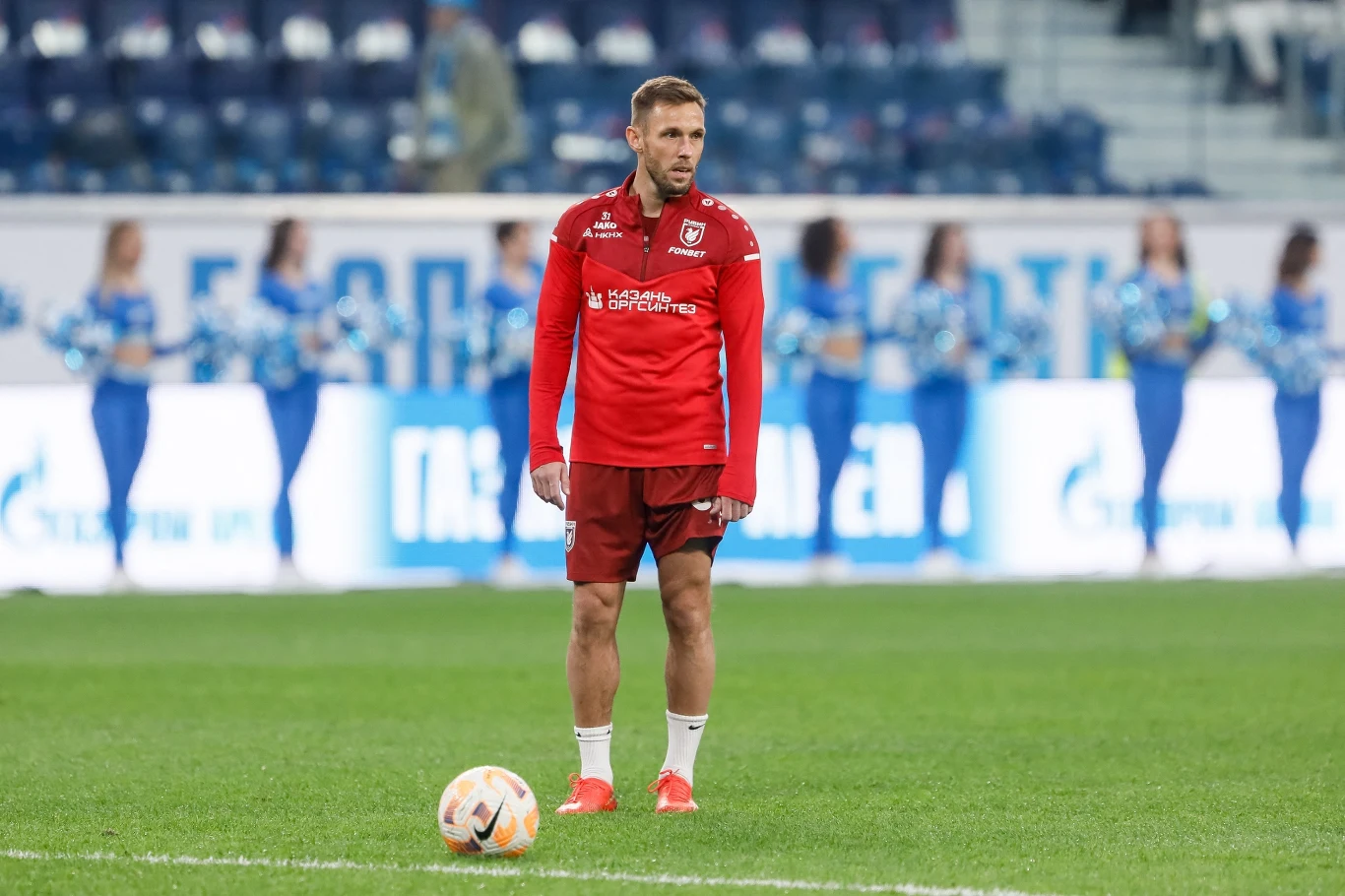 Piłkarz w czerwonym stroju sportowym stoi na murawie boiska, przodem do piłki nożnej, w tle widoczne są postacie cheerleaderek oraz trybuny stadionu.