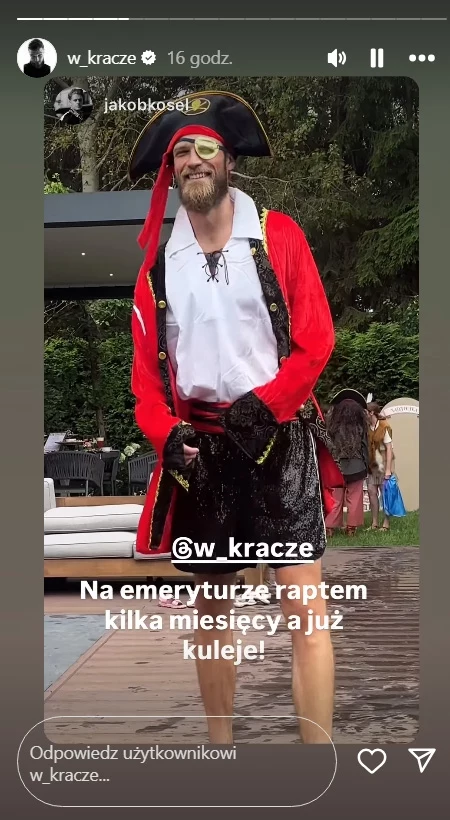 Andrzej Wrona przebrany za Kapitana Haka Mężczyzna przebrany za pirata z opaską na oku, kapeluszem i czerwoną bluzą stoi na zewnątrz, uśmiecha się, w tle widoczne osoby i zieleń.