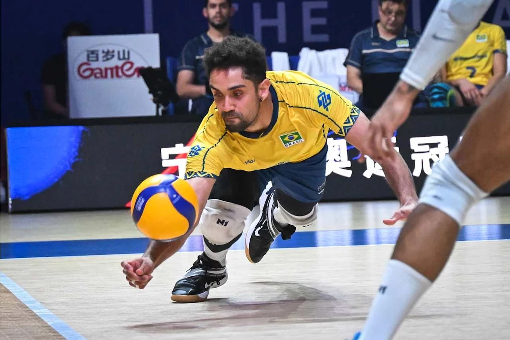 Zawodnik w żółtej koszulce reprezentacji Brazylii podczas dynamicznej próby obrony piłki do siatkówki, otoczony innymi graczami i trenerami na tle sportowej areny.