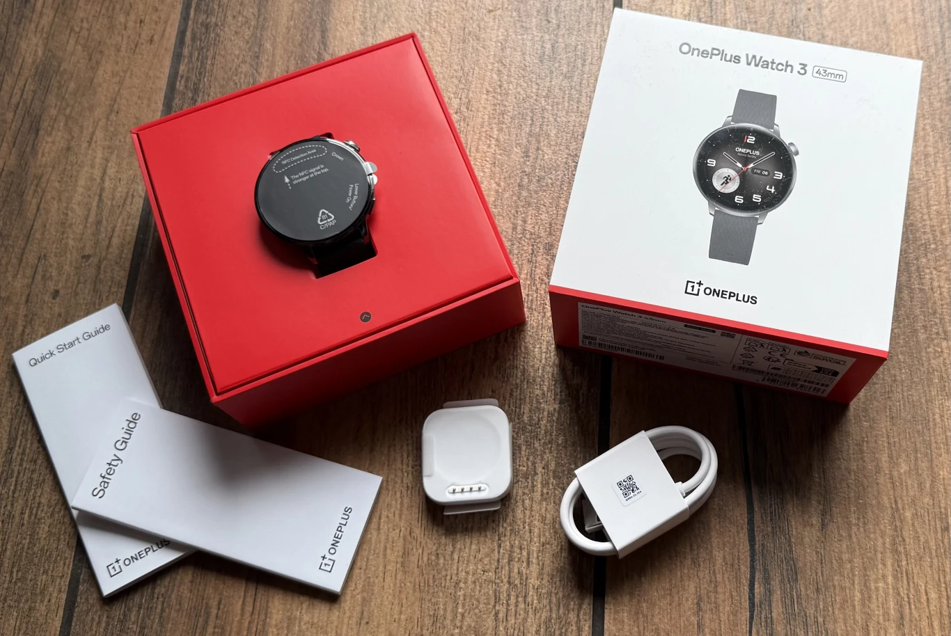 Zawartość opakowania z zegarkiem OnePlus Watch 3 43 mm. Smartwatch OnePlus Watch 3 w czerwonym pudełku, obok oryginalne opakowanie, przewód do ładowania, ładowarka indukcyjna oraz instrukcje obsługi umieszczone na drewnianej powierzchni.