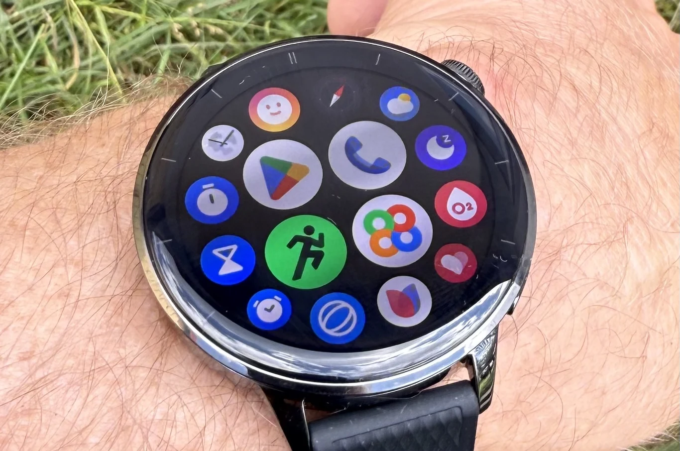 OnePlus Watch 3 pracuje pod kontrolą Wear OS i daje dostęp do wielu aplikacji Google. Inteligentny zegarek na nadgarstku, z wyświetlaczem prezentującym interfejs aplikacji w postaci kolorowych ikon na czarnym tle, na tarczy widoczne różne aplikacje, pasek zegarka wykonany z czarnego materiału, w tle fragment skóry i trawy.