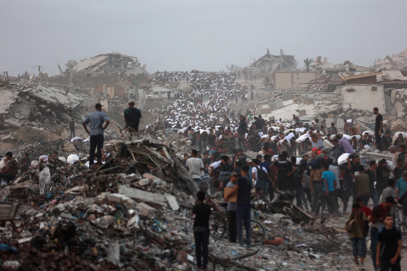 Palestyńczycy w drodze po pomoc humanitarną, desperacko próbują zdobyć mąkę Tłum osób przemieszcza się przez rozległe gruzowisko wśród zniszczonych budynków, otoczenie spowite kurzem i ruiny dominują w krajobrazie, ukazując skalę zniszczeń.