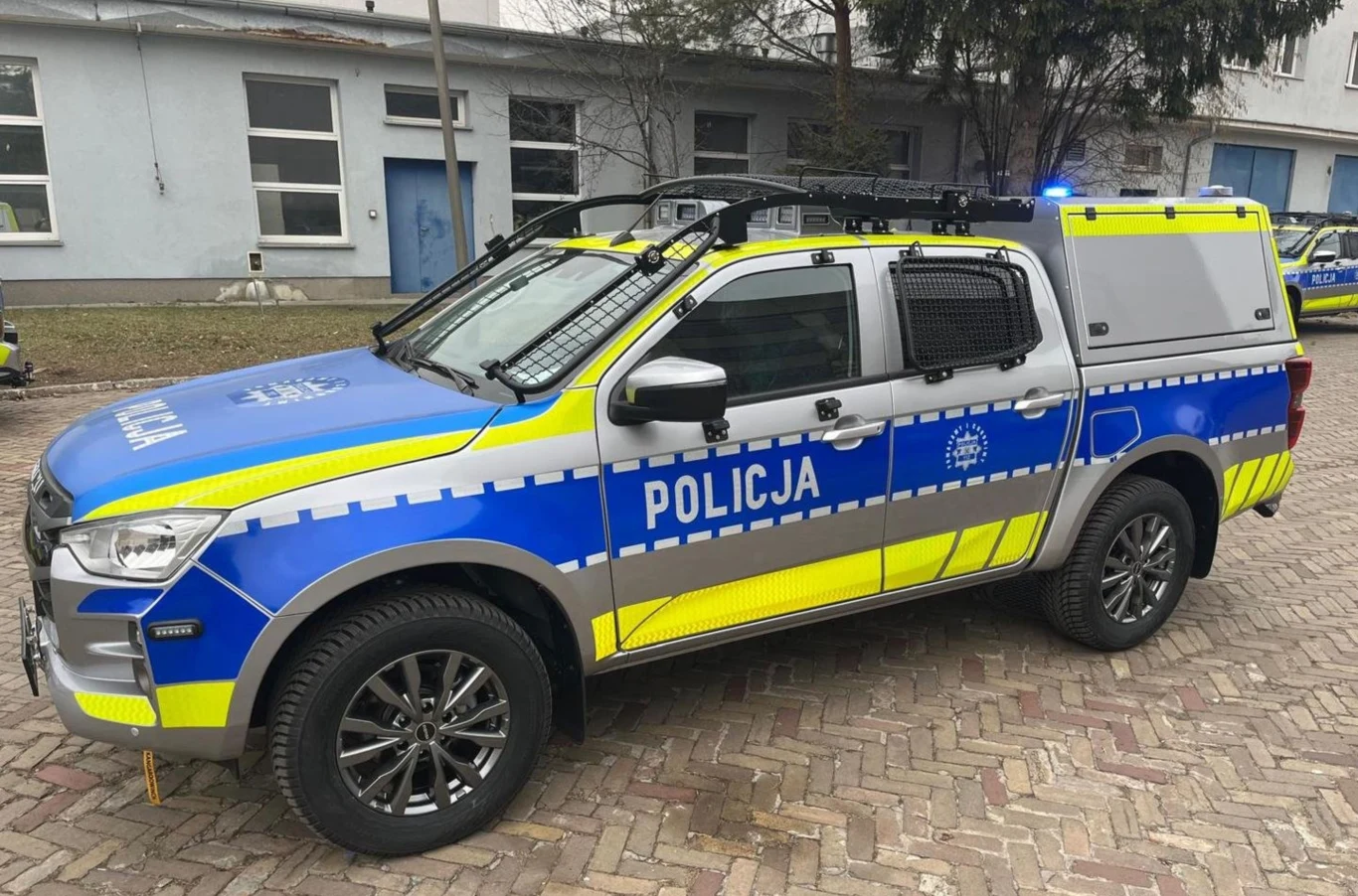 Policja będzie miała nowe radiowozy terenowe. Jakie? Nowoczesny policyjny samochód terenowy w kolorze niebieskim z elementami odblaskowymi i napisami POLICJA zatrzymany na brukowanej nawierzchni przed budynkiem służbowym.