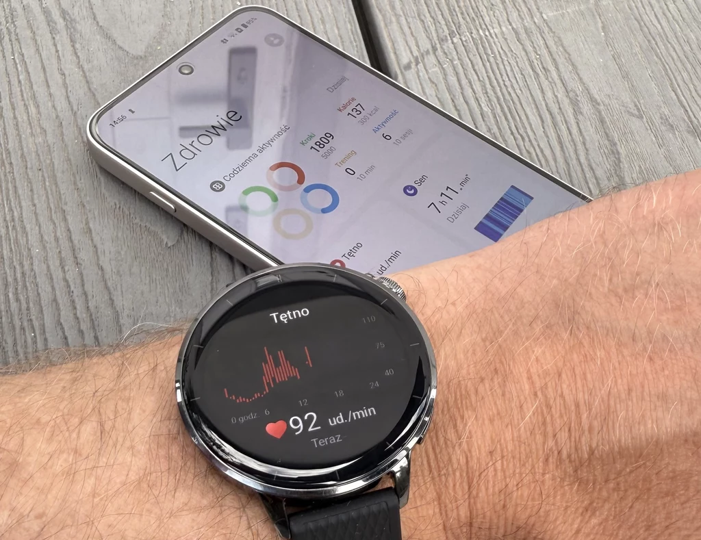 Smartwatch OnePlus Watch 3 oferuje wszystkie typowe funkcje z zakresu śledzenia kondycji i zdrowia użytkowników. Smartwatch na nadgarstku mierzący tętno, na ekranie wyświetla się wynik 92 uderzeń na minutę, w tle leży smartfon z uruchomioną aplikacją zdrowotną, która prezentuje dane o aktywności fizycznej użytkownika.