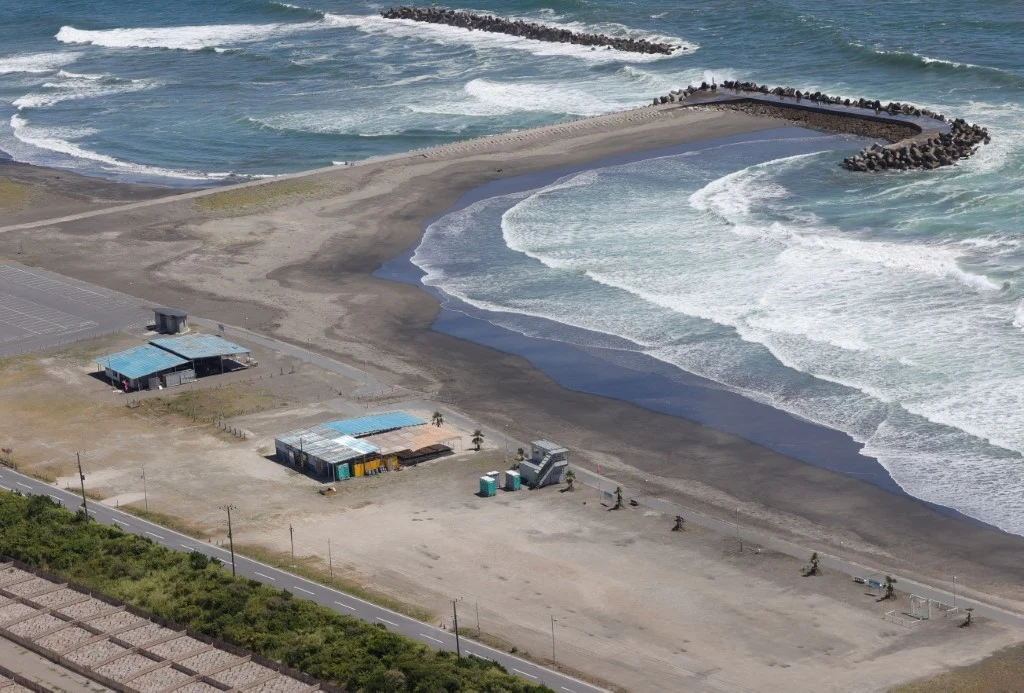 Plaża w prefekturze Chiba, Japonia Plaża z dwoma budynkami technicznymi i infrastrukturą publiczną, z falochronem oddzielającym część morza od brzegu, otoczona roślinnością i nadmorską drogą