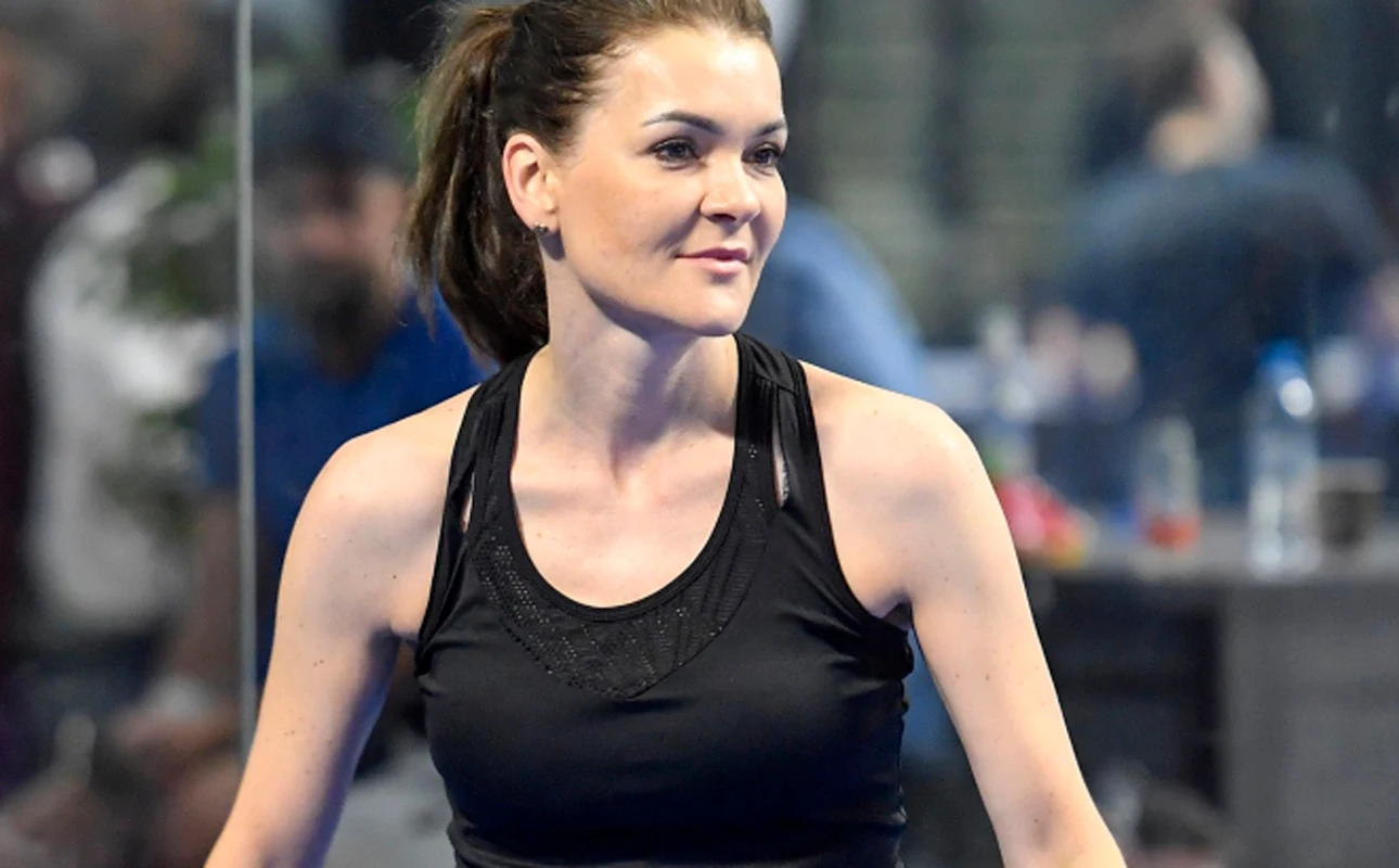 Kobieta w czarnej sportowej koszulce na tle rozmytego wnętrza, sportowy charakter sceny, skupiony wyraz twarzy, widoczne detale ubioru sportowego.