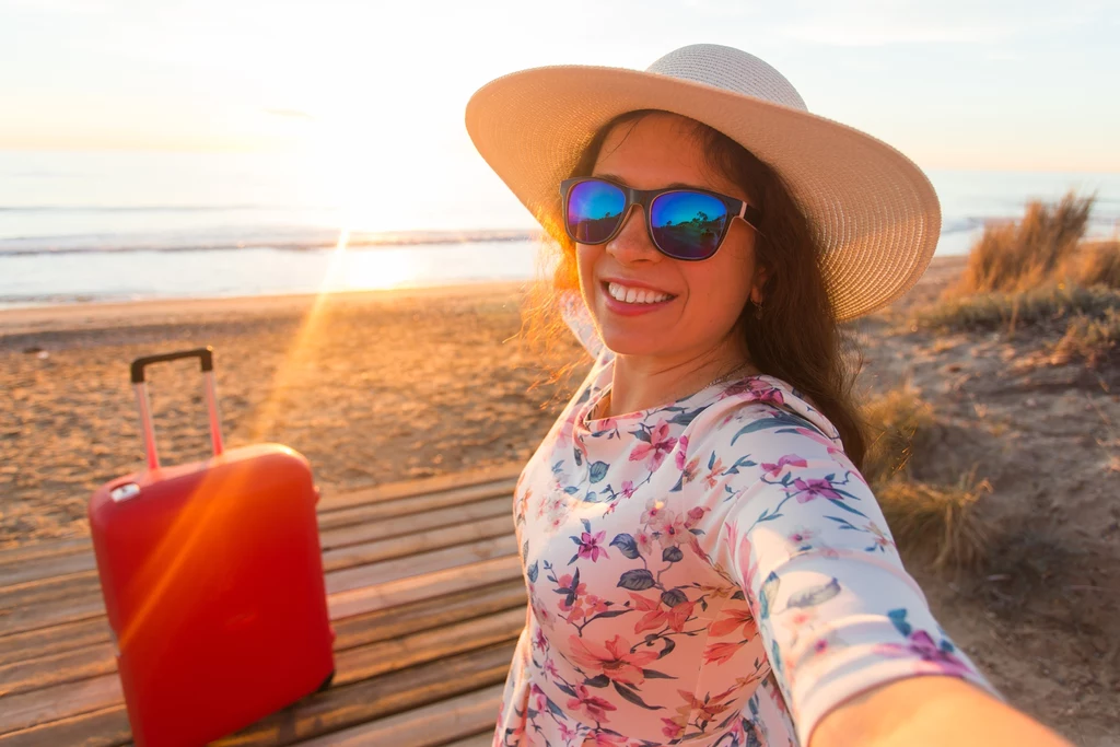 Trendy wakacyjne Uśmiechnięta kobieta w okularach przeciwsłonecznych i kapeluszu robi selfie na plaży przy zachodzie słońca, obok niej czerwona walizka.