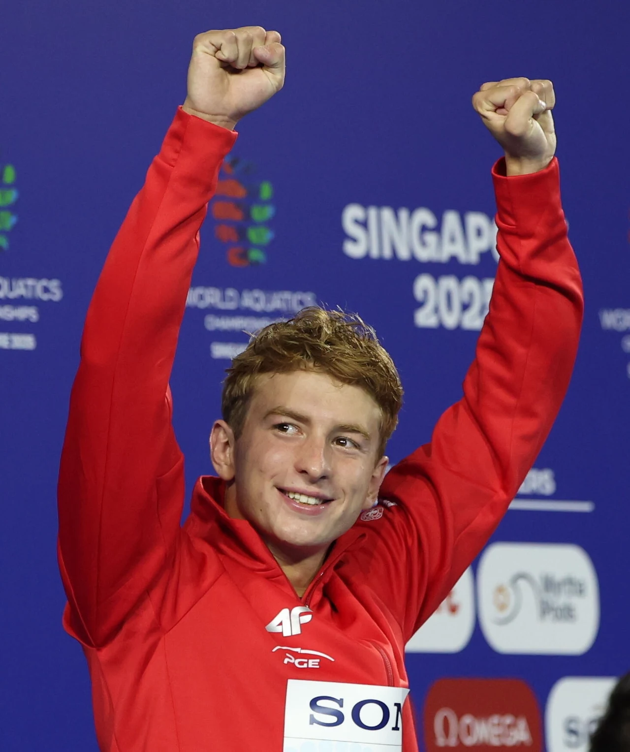 Krzysztof Chmielewski, Singapur 2025 Sportowiec w czerwonej bluzie unosi ręce w geście triumfu na tle niebieskiej ścianki z napisem 'Singapur 2024' oraz logotypami wydarzenia sportowego.