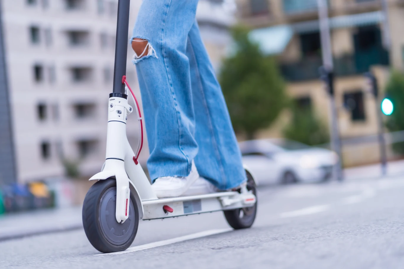 Osoba w niebieskich, lekko podartych dżinsach i białych sportowych butach jedzie białą hulajnogą elektryczną po miejskiej drodze, w tle rozmyte budynki i samochody.
