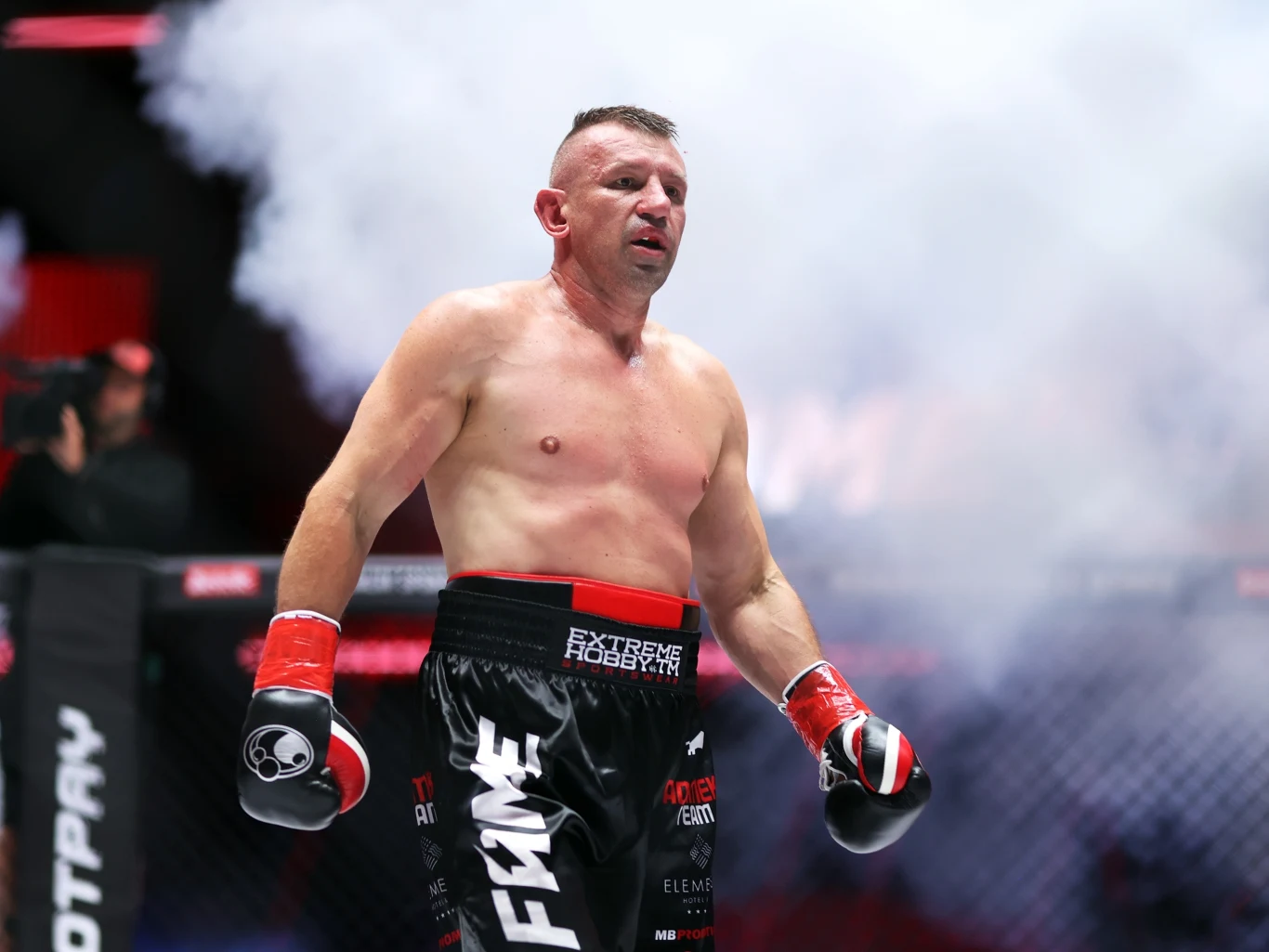 Tomasz Adamek w FAME Muskularny mężczyzna w sportowym stroju MMA stoi na ringu, wokół niego unoszą się kłęby dymu, w tle widoczne fragmenty ogrodzenia i oświetlenie areny.
