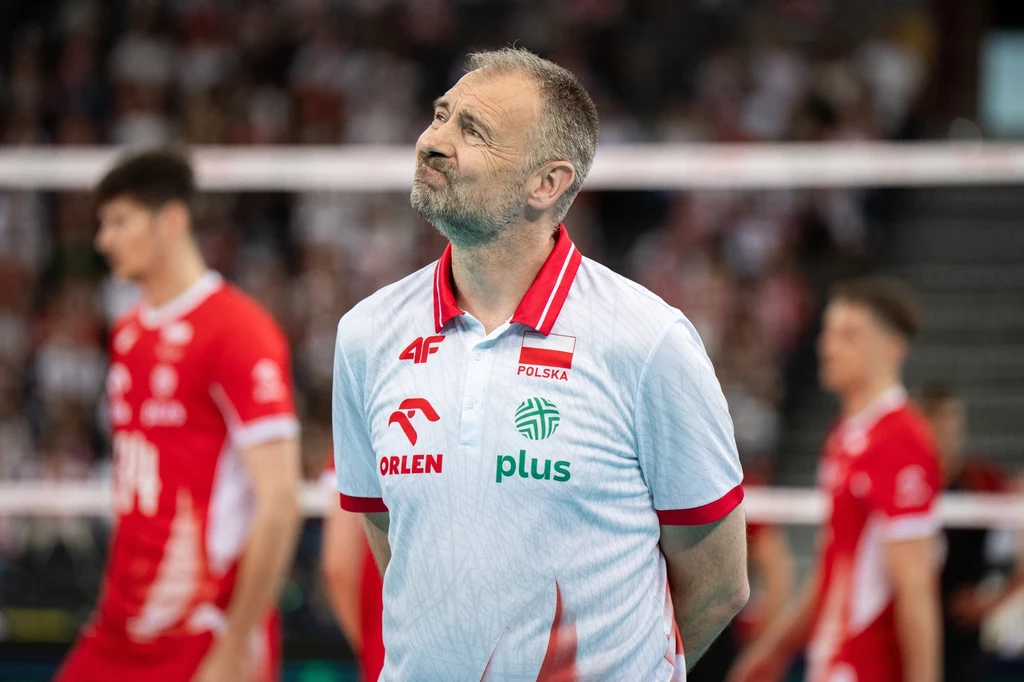 Nikola Grbić Trener reprezentacji Polski w siatkówce w białej koszulce z logo sponsorów, wyrażający emocje, w tle rozmazani zawodnicy w czerwonych strojach siatkarskich.