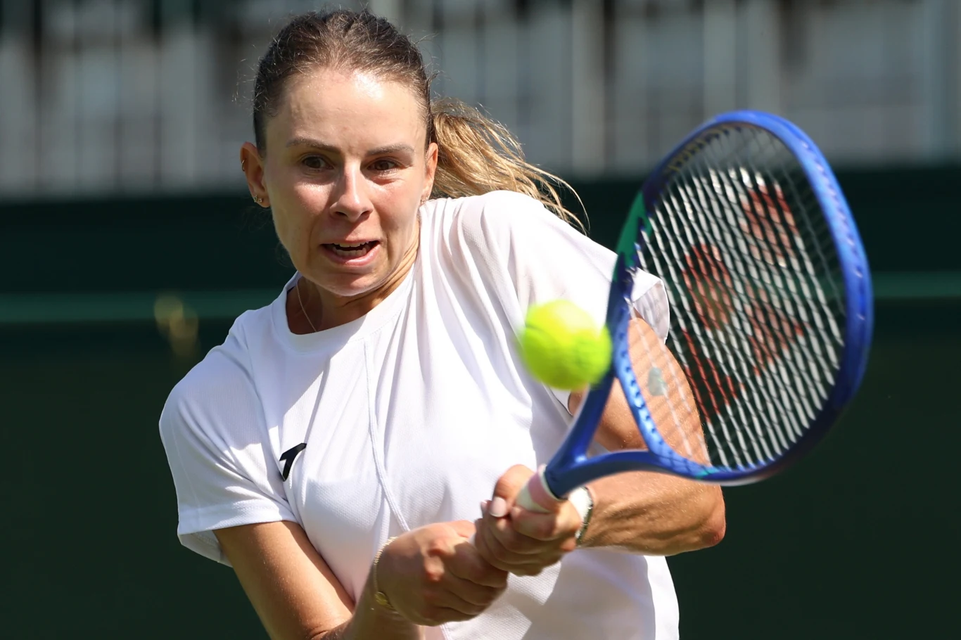 Magda Linette Kobieta w białej koszulce wykonuje zamach na piłkę tenisową dwuręcznym backhandem, trzymając niebieską rakietę tenisową. W tle znajdują się elementy kortu tenisowego.