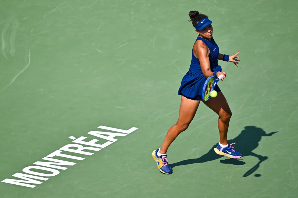 Naomi Osaka, Montreal 2025 Tenisistka podczas gry na korcie, uderza piłkę rakietą, ubrana w niebieską sportową sukienkę i czapkę, na zielonej nawierzchni z napisem 'Montréal', jasne oświetlenie i ruch sportowy.