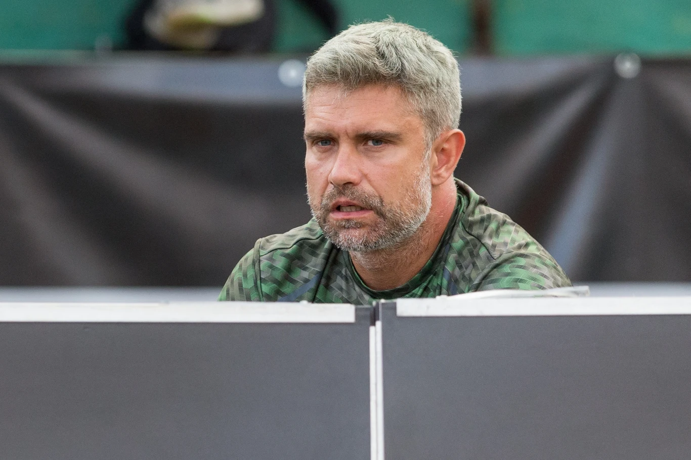 Tomasz Wiktorowski Mężczyzna w średnim wieku o siwych włosach i brodzie siedzi za stołem, ubrany w koszulkę w odcieniach zieleni, tło rozmyte z ciemnym banerem.