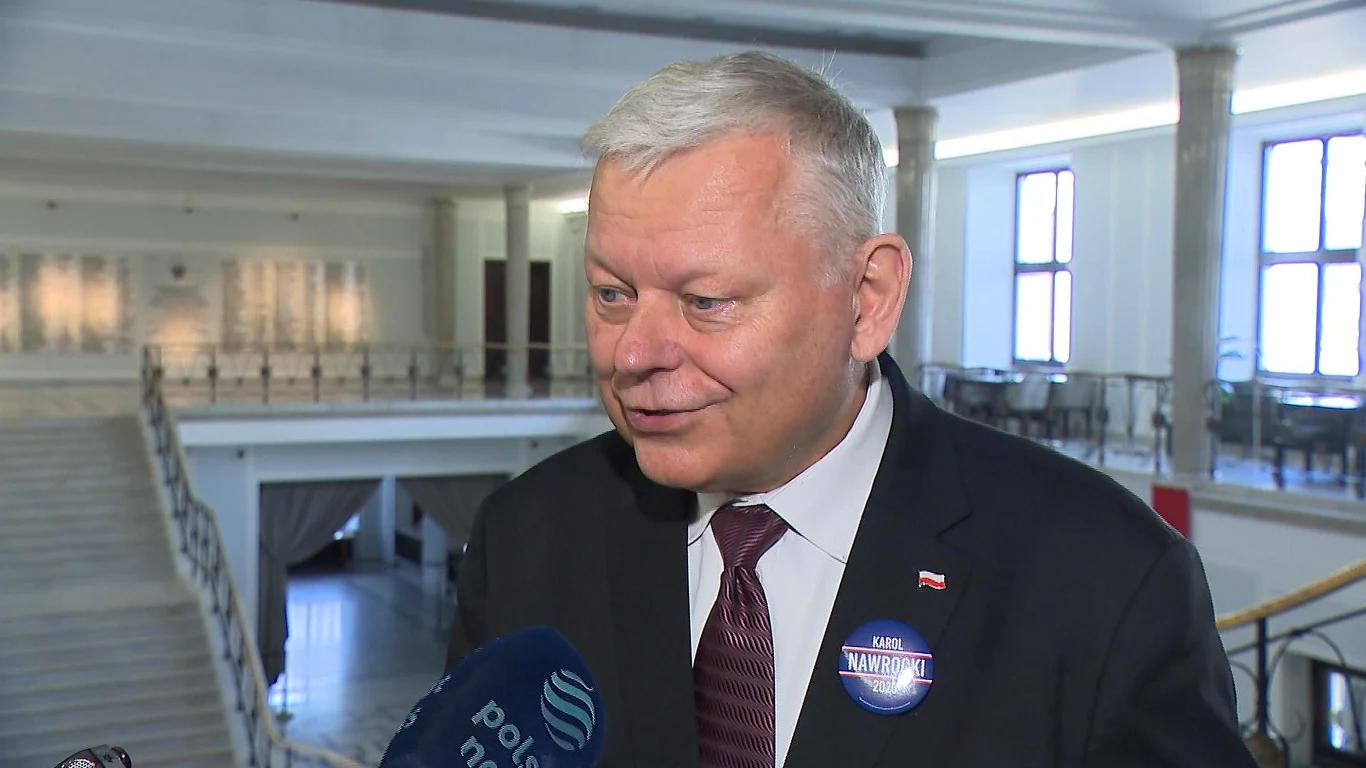 Jest śledztwo dotyczące Marka Suskiego, polityka PiS Mężczyzna w garniturze z przypiętym identyfikatorem oraz mikrofon z logo policji w przestronnym, jasnym wnętrzu budynku administracyjnego lub urzędowego.