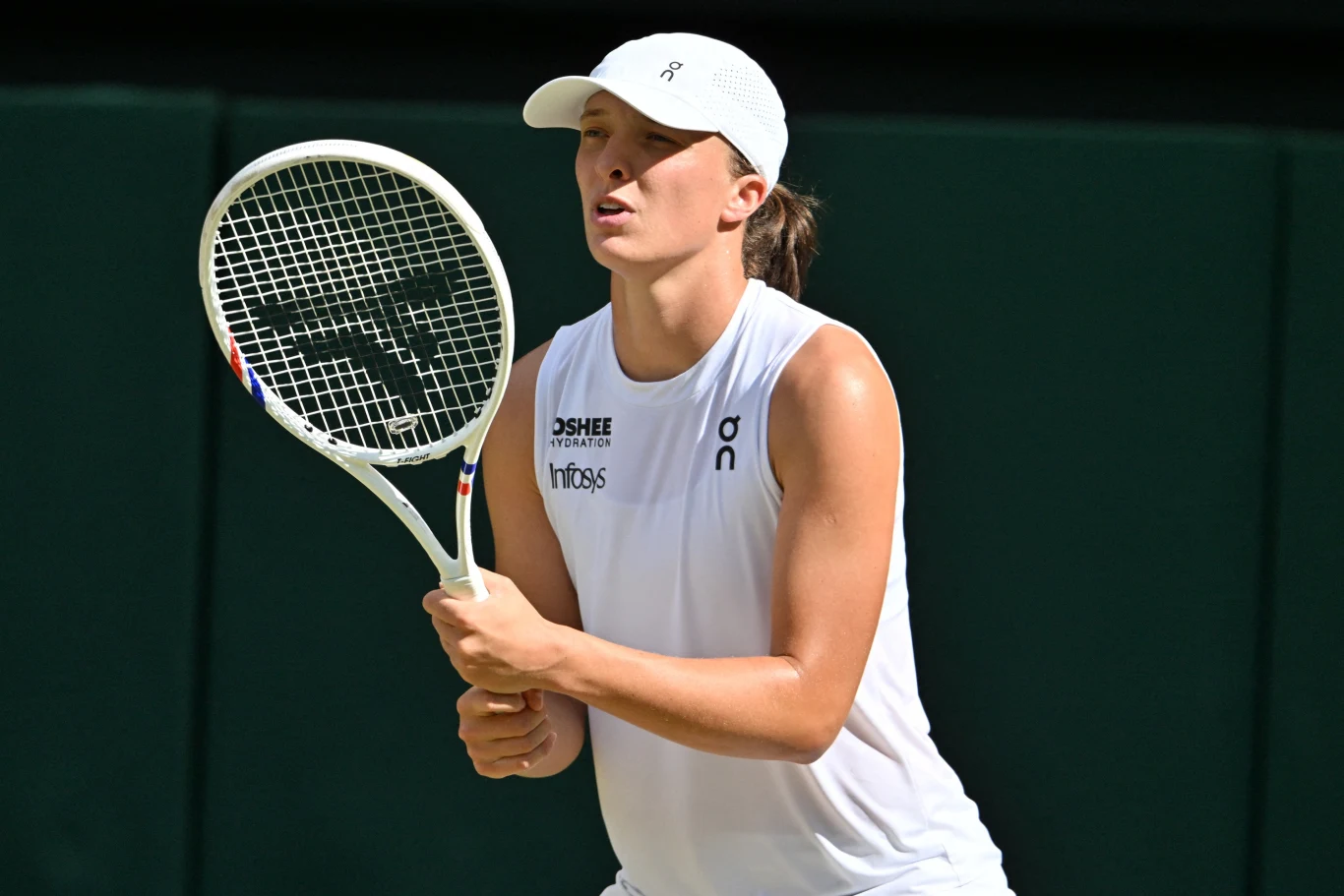 Iga Świątek rywalizowała z Hanyu Guo o awans do trzeciej rundy WTA 1000 w Montrealu Tenisistka w białym stroju sportowym i czapce z daszkiem przygotowuje się do odbioru piłki podczas meczu, trzymając oburącz rakietę tenisową na tle ciemnego kortu.
