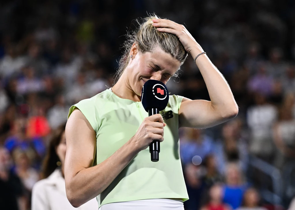 Eugenie Bouchard Kobieta w jasnej koszulce, trzymająca mikrofon jedną ręką, a drugą poprawiająca włosy, wyraża silne emocje; w tle rozmyta publiczność podczas wydarzenia sportowego.