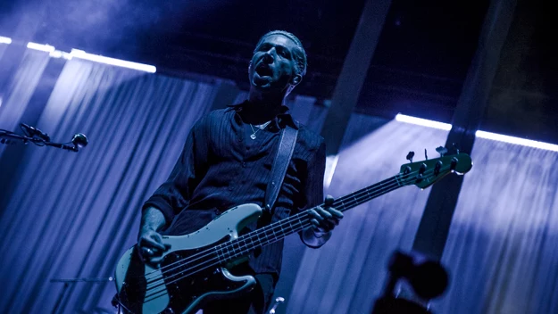 Muzyk grający na gitarze basowej na scenie koncertowej oświetlonej niebieskim światłem, z kurtynami w tle oraz widoczną dynamiczną atmosferą występu na żywo