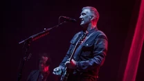 Muzyk stoi na scenie w świetle czerwonych reflektorów, śpiewa do mikrofonu i gra na gitarze elektrycznej, w tle drugi gitarzysta jest słabo widoczny w cieniu.